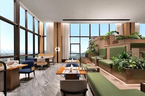 En modern hotellobby med stora fönster, sittgrupper och en vägg täckt av gröna växter.