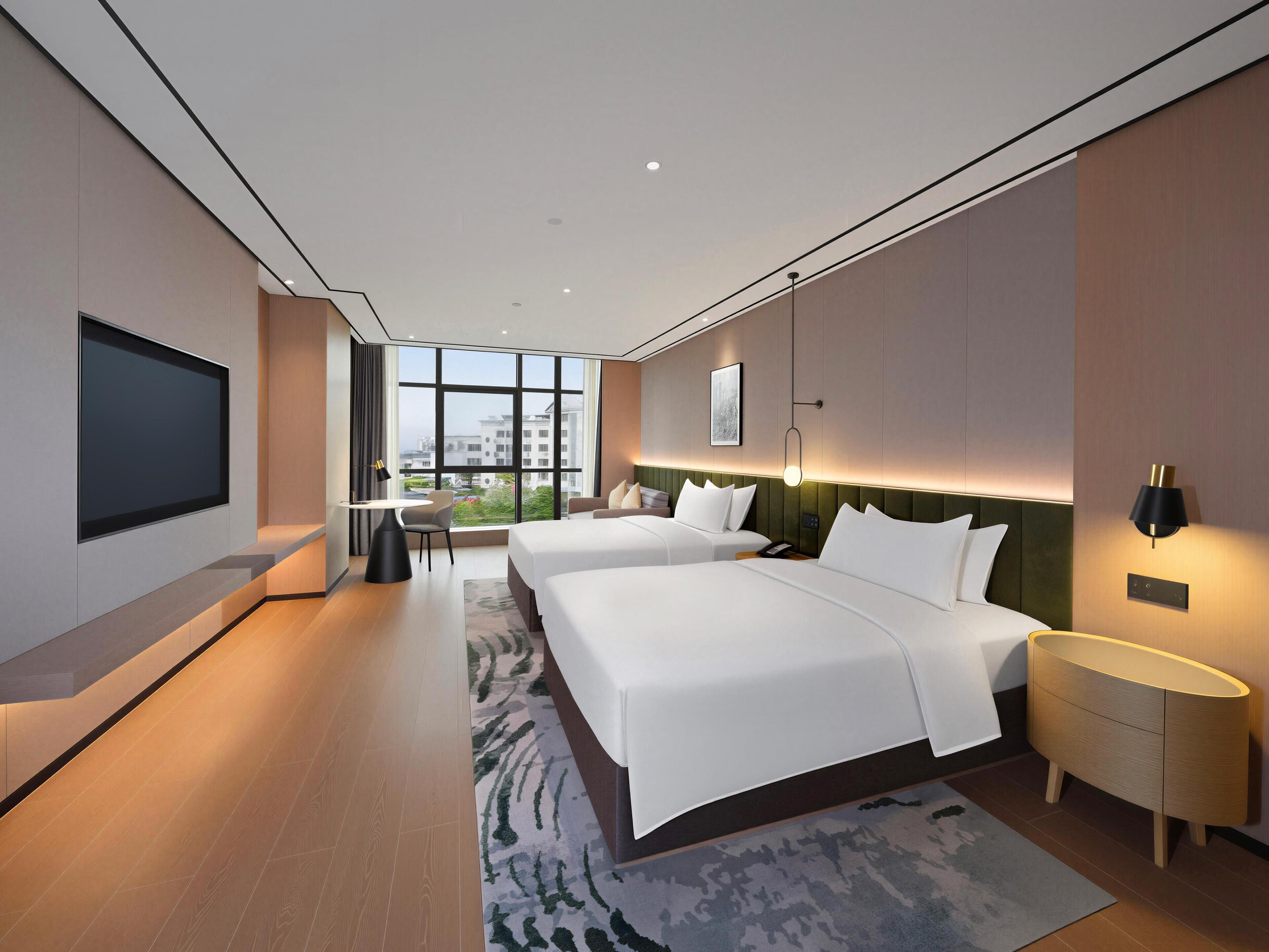 A modern hotel room with two beds, a flat-screen TV, a desk, and a large window offering a city view.