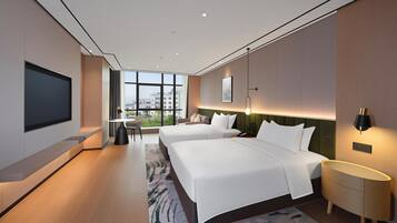 A modern hotel room with two beds, a flat-screen TV, a desk, and a large window offering a city view.