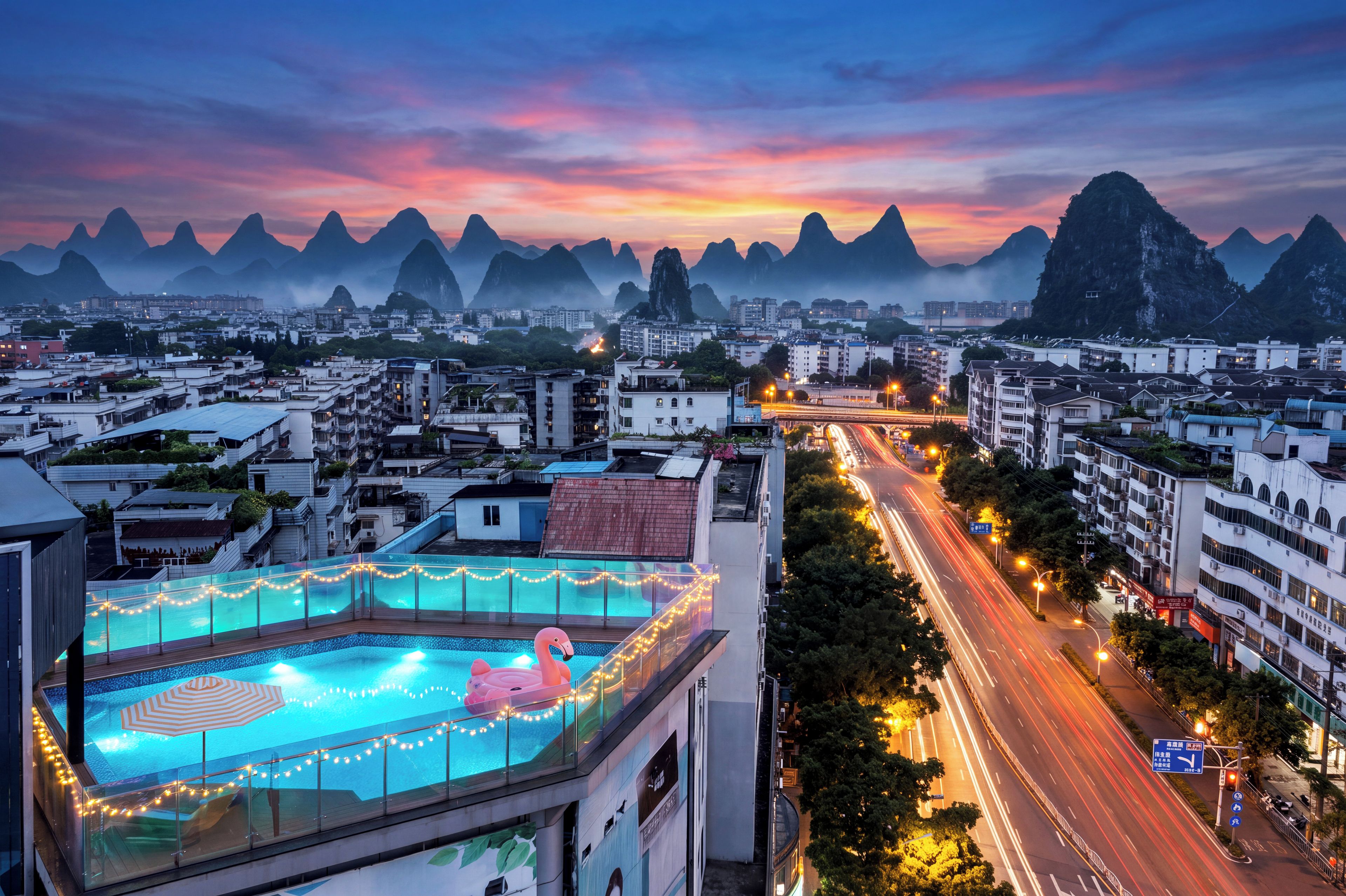A cityscape with a pool area featuring a flamingo float and string lights, surrounded by buildings and a road with light trails.