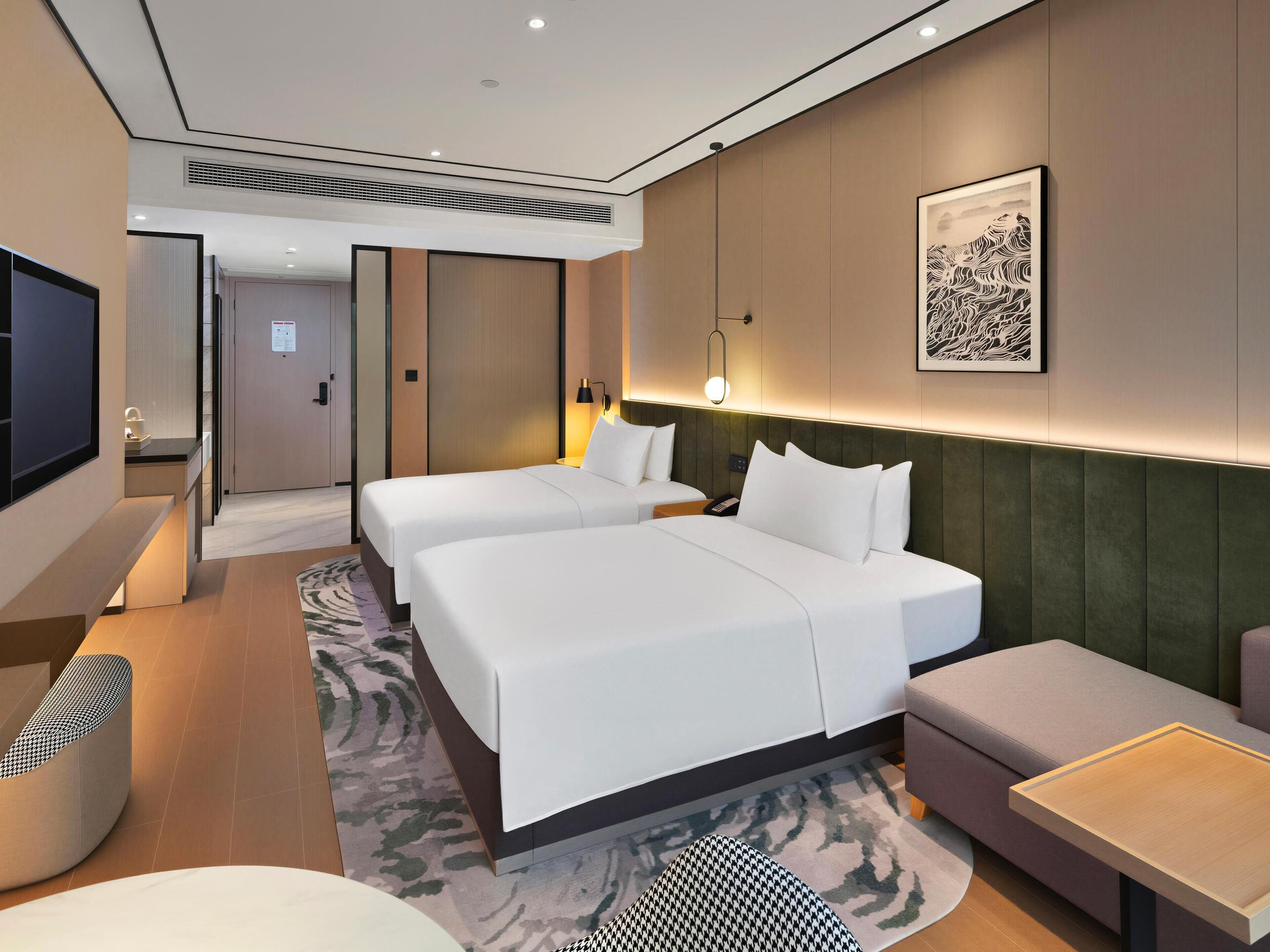 A hotel room with two beds, a sofa, a small table, a TV, and a framed artwork on the wall.