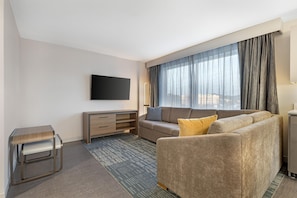 A modern hotel room with a sofa, a flat-screen TV mounted on the wall, a bedside table, and a window with curtains. - Homewood Suites by Hilton Walker Grand Rapids North (Grand Rapids)