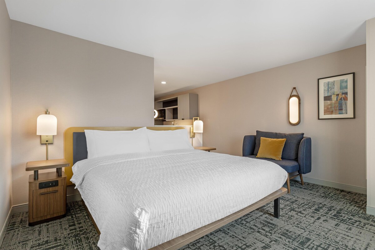 A hotel room with a large bed, bedside lamps, a small table, a sofa, and a painting on the wall.