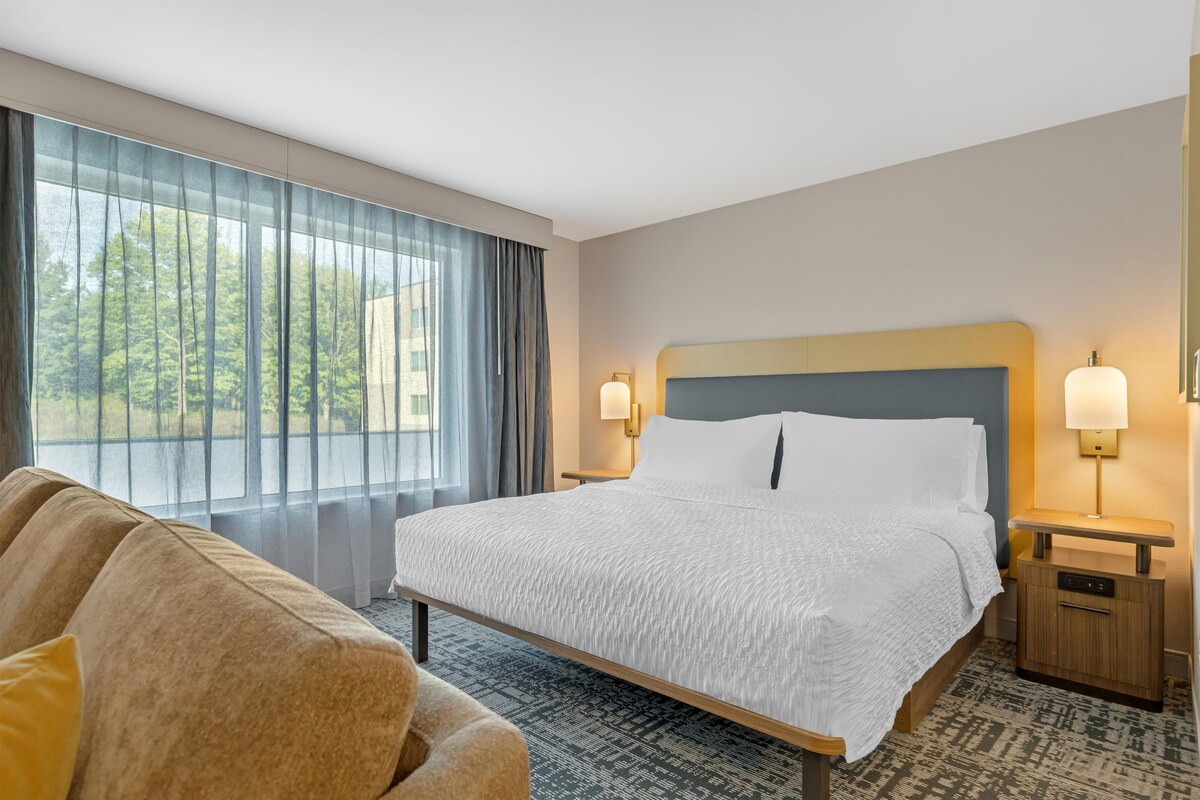 A hotel room with a large bed, two bedside lamps, a sofa, and a view of trees through the window.