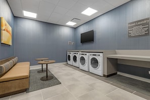 A laundry room with a sofa, two small tables, a flat-screen TV, and a washing machine. - Homewood Suites by Hilton Walker Grand Rapids North (Grand Rapids)