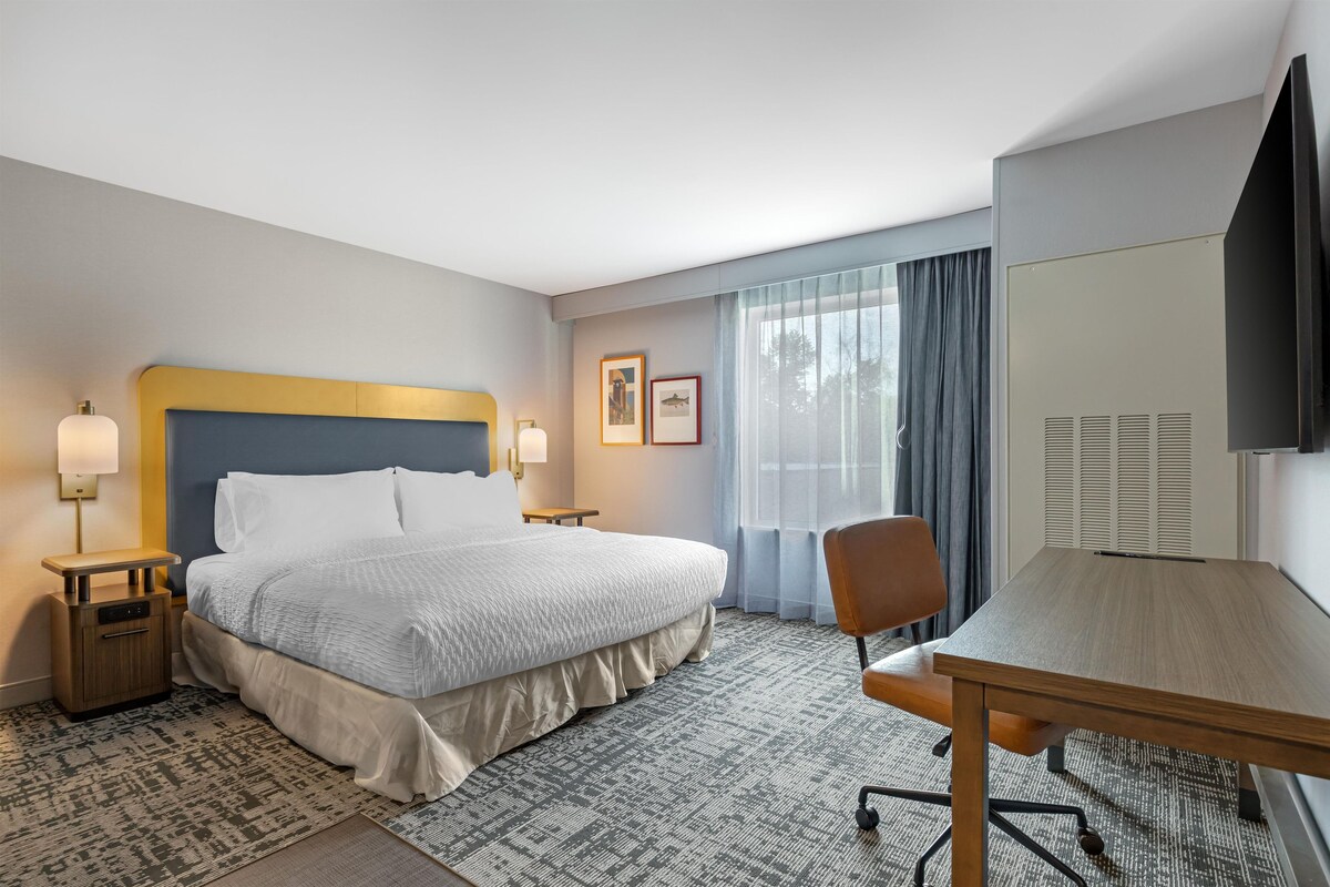 A hotel room with a bed, bedside tables, a desk, a chair, a large window with curtains, and a flat-screen TV.