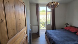 Une chambre à coucher avec un lit, une armoire en bois, un radiateur, un chauffage d’appoint, un luminaire au plafond et une vue sur l’extérieur, à travers une porte ouverte.