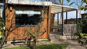 Une cabane en bois moderne dotée d’une grande fenêtre, d’un patio avec une table et des chaises, et d’un petit arbre au premier plan.