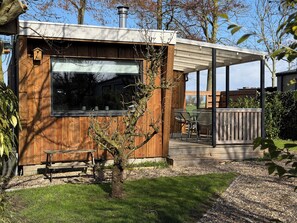 Eine moderne Holzhütte mit großem Fenster, einer Terrasse mit Tisch und Stühlen und einem kleinen Baum im Vordergrund.