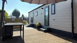 A deck with a table and chairs, a patio umbrella, and a mobile home with large windows and a small garden. - Beautiful new chalet with private parking space on the edge of the park. (Sint Maarten)