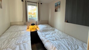 A room with two beds, a window with curtains, a radiator, and a painting on the wall. - Beautiful new chalet with private parking space on the edge of the park. (Sint Maarten)