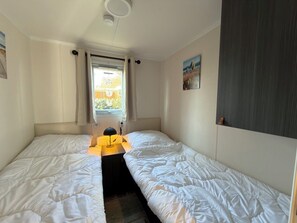 A room with two beds, a window with curtains, a radiator, and a painting on the wall. - Beautiful new chalet with private parking space on the edge of the park. (Sint Maarten)