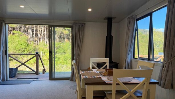 A dining area with a table and chairs, a view of greenery outside, and a chimney. - Alpaca Farm Retreat - Peaceful Rural Escape (Lower Hutt)