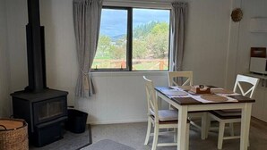 A room with a wood-burning stove, a dining table with chairs, a window with curtains, and a view of greenery outside. - Alpaca Farm Retreat - Peaceful Rural Escape (Lower Hutt)