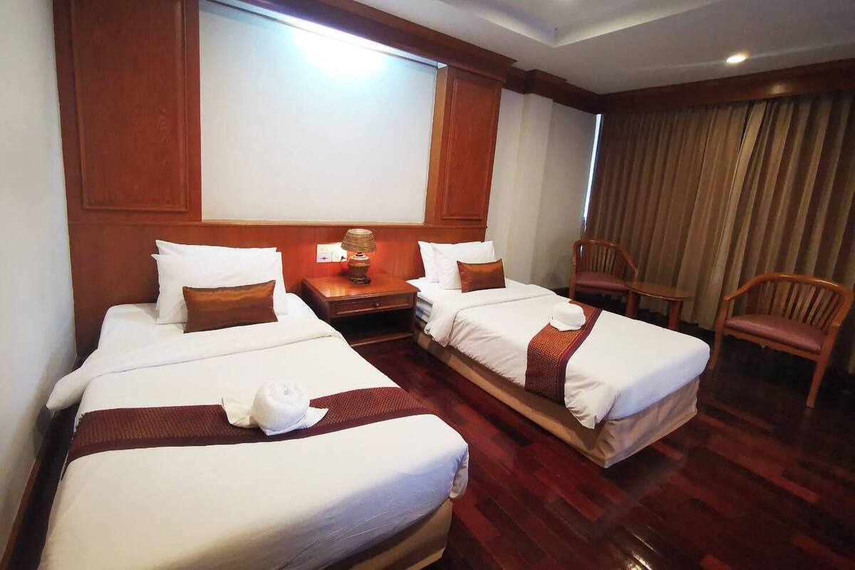 Una habitación de hotel con dos camas, una mesita de noche, una lámpara, una silla y un ventanal con cortinas.