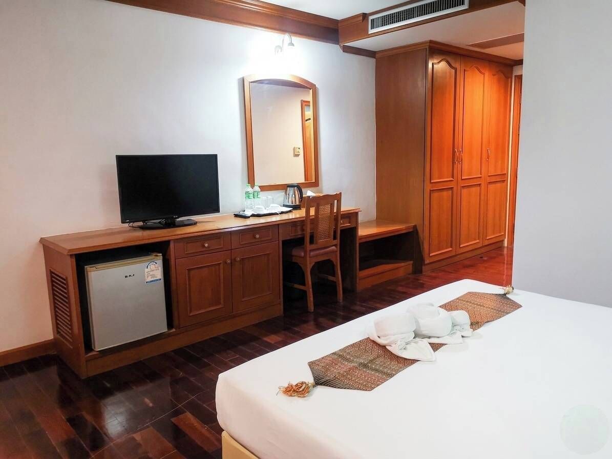 A hotel room with a bed, a desk with a television, a mirror, a wardrobe, and a small refrigerator.