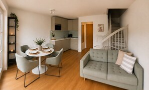 Ein modernes Wohnzimmer mit einem grauen Sofa, einem Esstisch für zwei Personen und einer Küche im Hintergrund.