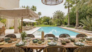 Zona pranzo a bordo piscina con tavolo apparecchiato, poltroncine da relax e ombrellone di paglia.