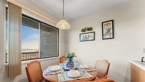 Uma sala de jantar com uma mesa redonda, quatro cadeiras, um vaso com flores e vista para um corpo d'água por uma grande janela.