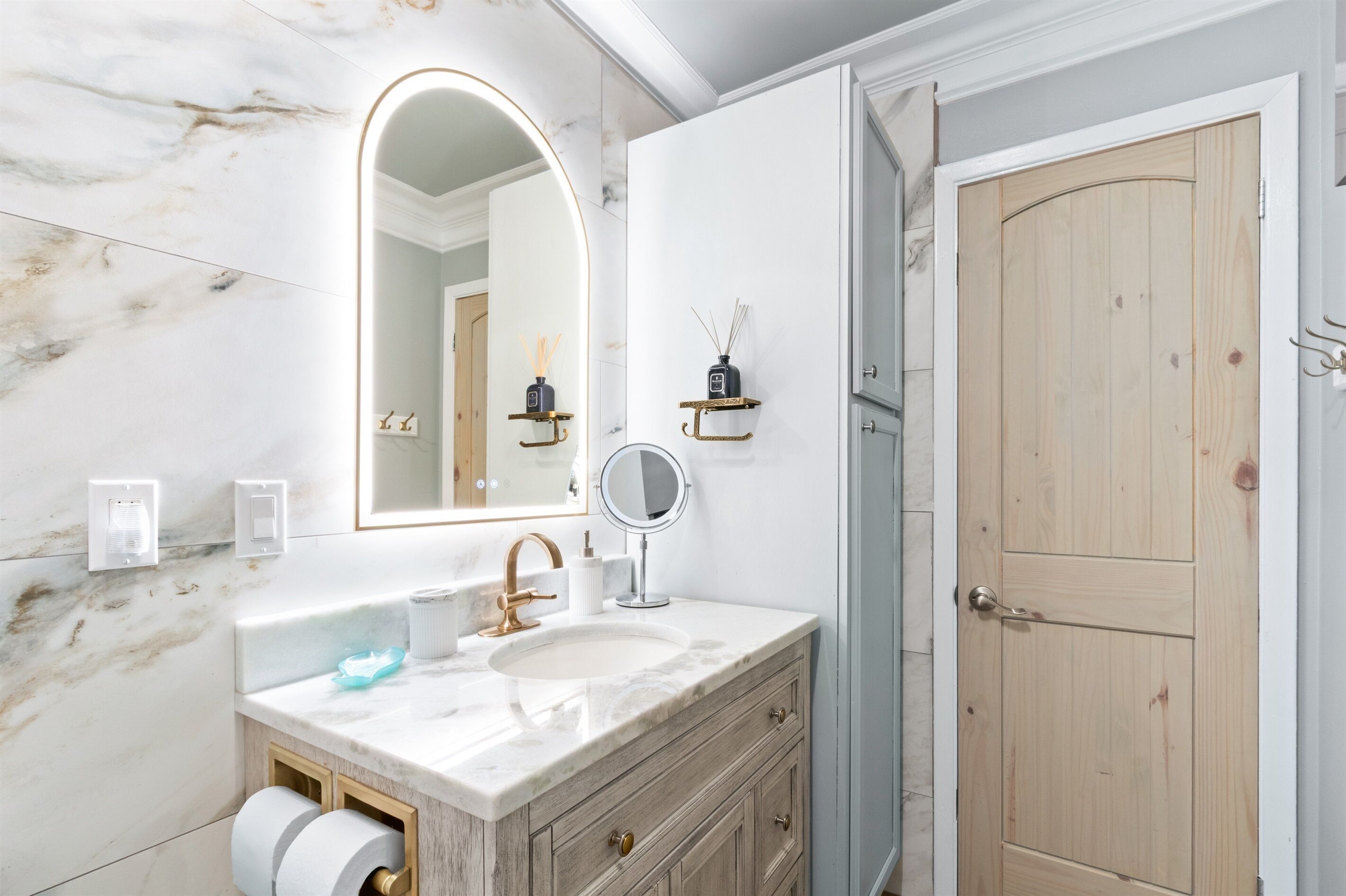 Une salle de bain avec un meuble-lavabo en marbre, un grand miroir, une porte en bois et une petite étagère sur laquelle se trouvent un flacon et un diffuseur à tiges.
