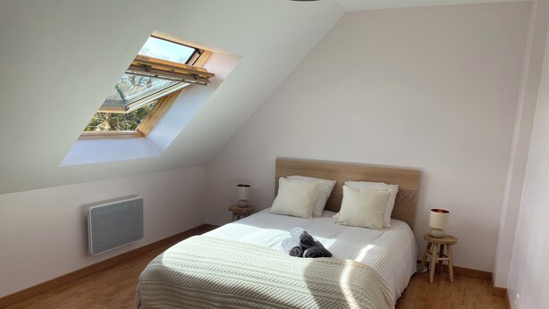 Un dormitorio con techo inclinado, tragaluz, cama con manta blanca, dos mesitas de noche con lámparas y un radiador.