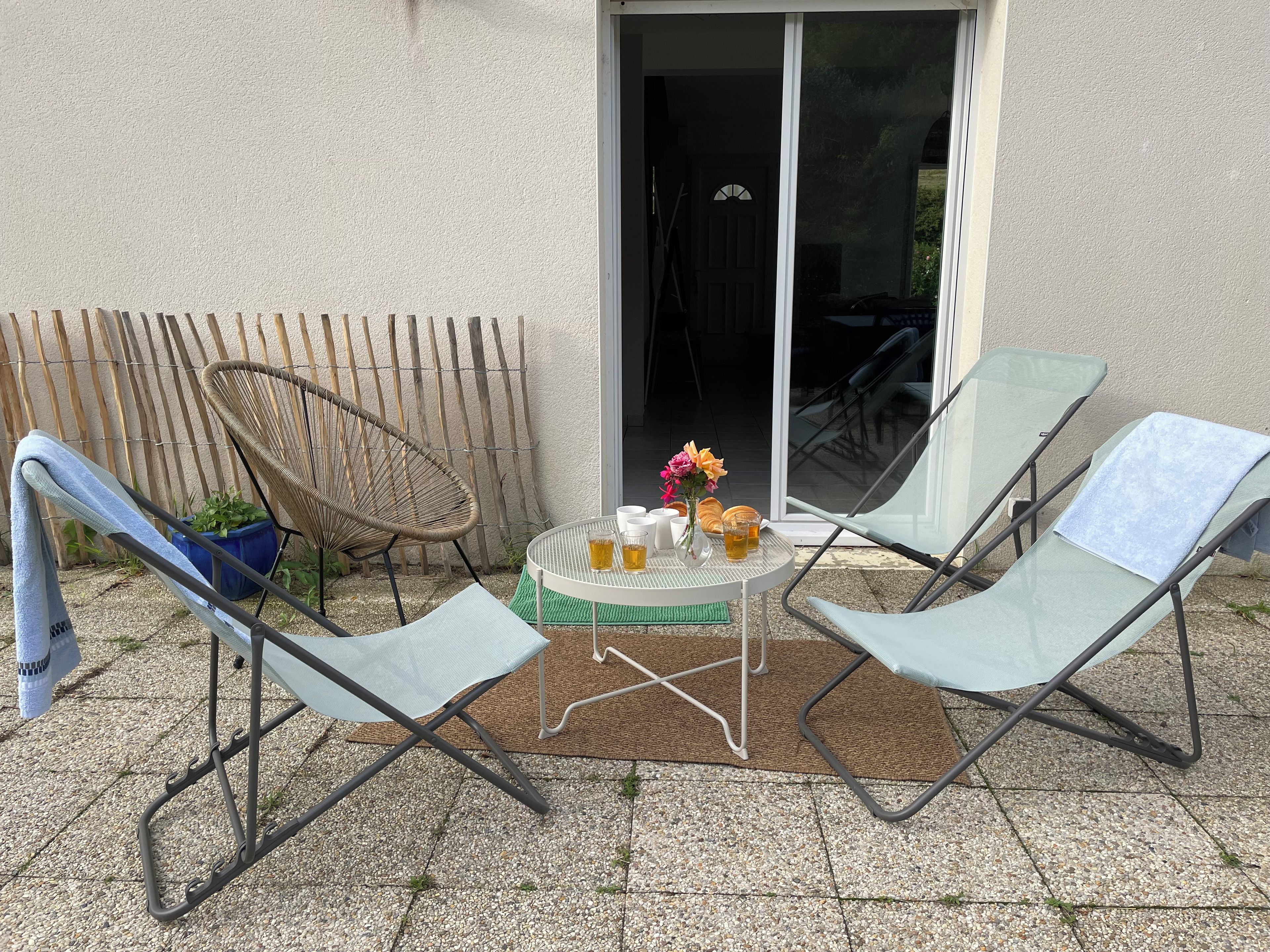 An outdoor seating area with two lounge chairs, a small round table with drinks and a vase of flowers, and a bamboo fence in the background.