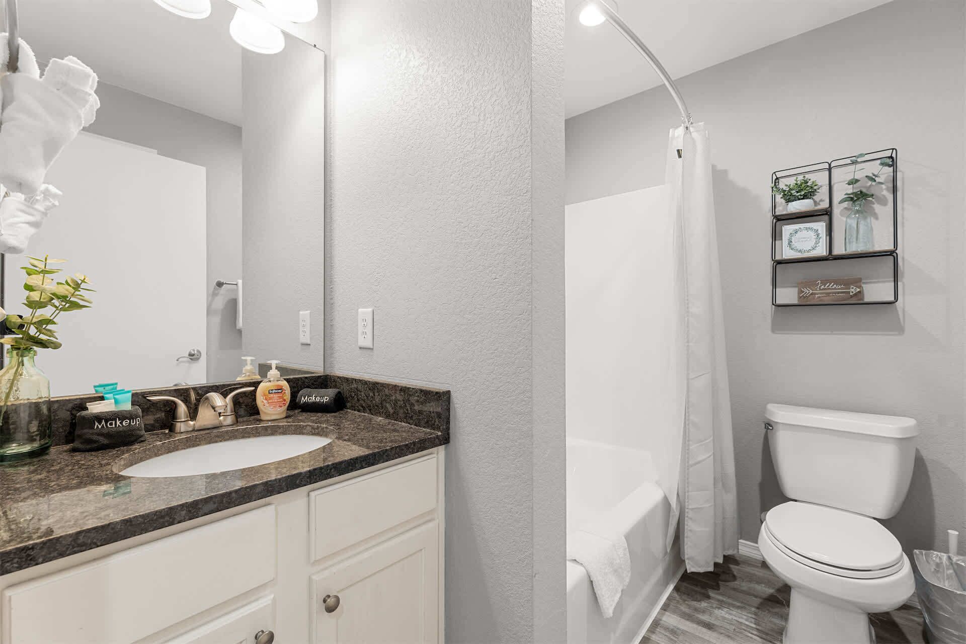 A bathroom with a white sink, a mirror, a shower curtain, a toilet, and a shelf with decorative items.