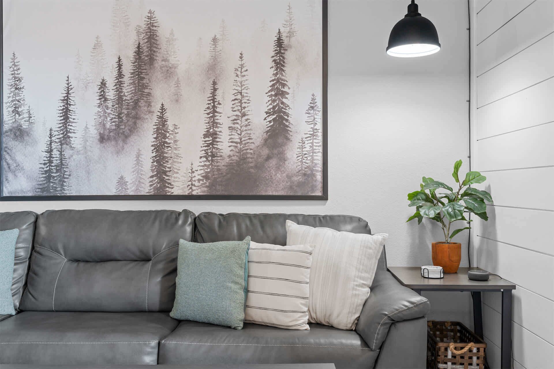 Ein graues Ledersofa mit Kissen, ein gerahmtes Bild eines Waldes, eine hängende Pendelleuchte und ein Blumentopf auf einem Beistelltisch.