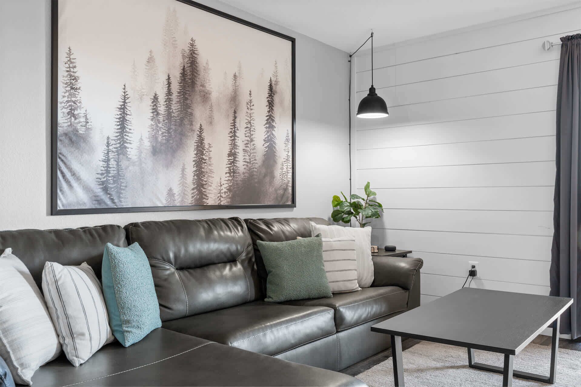 A modern living room with a leather sofa, a coffee table, a large forest-themed artwork, a pendant light, and a potted plant.