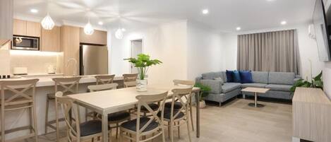 Ein modernes Wohnzimmer mit Essbereich, einem grauen Sofa, einem Flachbildfernseher und einer Küche mit Holzschränken und einem Edelstahlkühlschrank.