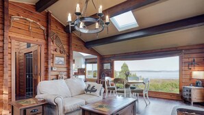 A cozy cabin interior with a wooden log wall, a sofa, a dining table with chairs, a chandelier, and a view of the outdoors. - Ocean View Guest House - stunning Ocean Views by Victoria Prime!! (Saanichton)
