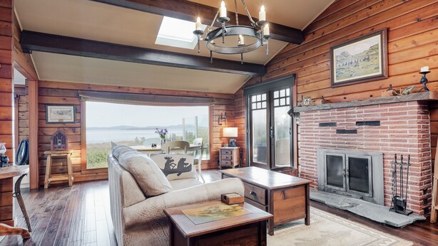 A log cabin interior with a fireplace, a sofa, a dining table, and a view of the outdoors. - Ocean View Guest House - stunning Ocean Views by Victoria Prime!! (Saanichton)