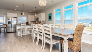 A modern kitchen with white cabinetry, a stainless steel refrigerator, and a dining area with white chairs and a wooden table. - Topsail Fairytale - with sweeping ocean views from three front decks! (Surf City)