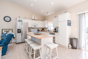 A modern kitchen with white cabinetry, a central island with stools, a refrigerator, microwave, and a coffee maker. - Sunsets and Sand- 4BR, Dog Friendly, Ocean Views (N. Topsail Beach)