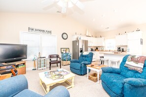 A living room with a TV, blue armchairs, a coffee table with magazines, and a kitchen in the background. - Sunsets and Sand- 4BR, Dog Friendly, Ocean Views (N. Topsail Beach)