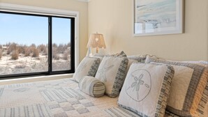 A bedroom with a large window, a bed with patterned bedding, a lamp, and a framed picture on the wall. - Oceanfront Dunes Condo w/ Beach Views & Access (Ocean City)