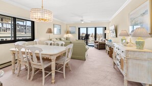 A spacious room with a dining table, chairs, a sofa, a cabinet, and a large window offering a view. - Oceanfront Dunes Condo w/ Beach Views & Access (Ocean City)
