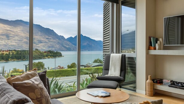 Una sala de estar moderna con un gran ventanal que da a un balcón con vistas a un lago y montañas.