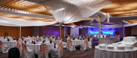 Une salle de banquet avec des tables rondes recouvertes de nappes blanches, des chaises ornées de rubans orange et une scène éclairée de lumière bleue.