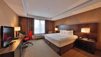 A hotel room with a large bed, a desk with a TV, a chair, a bedside table with a lamp, and a window with curtains.