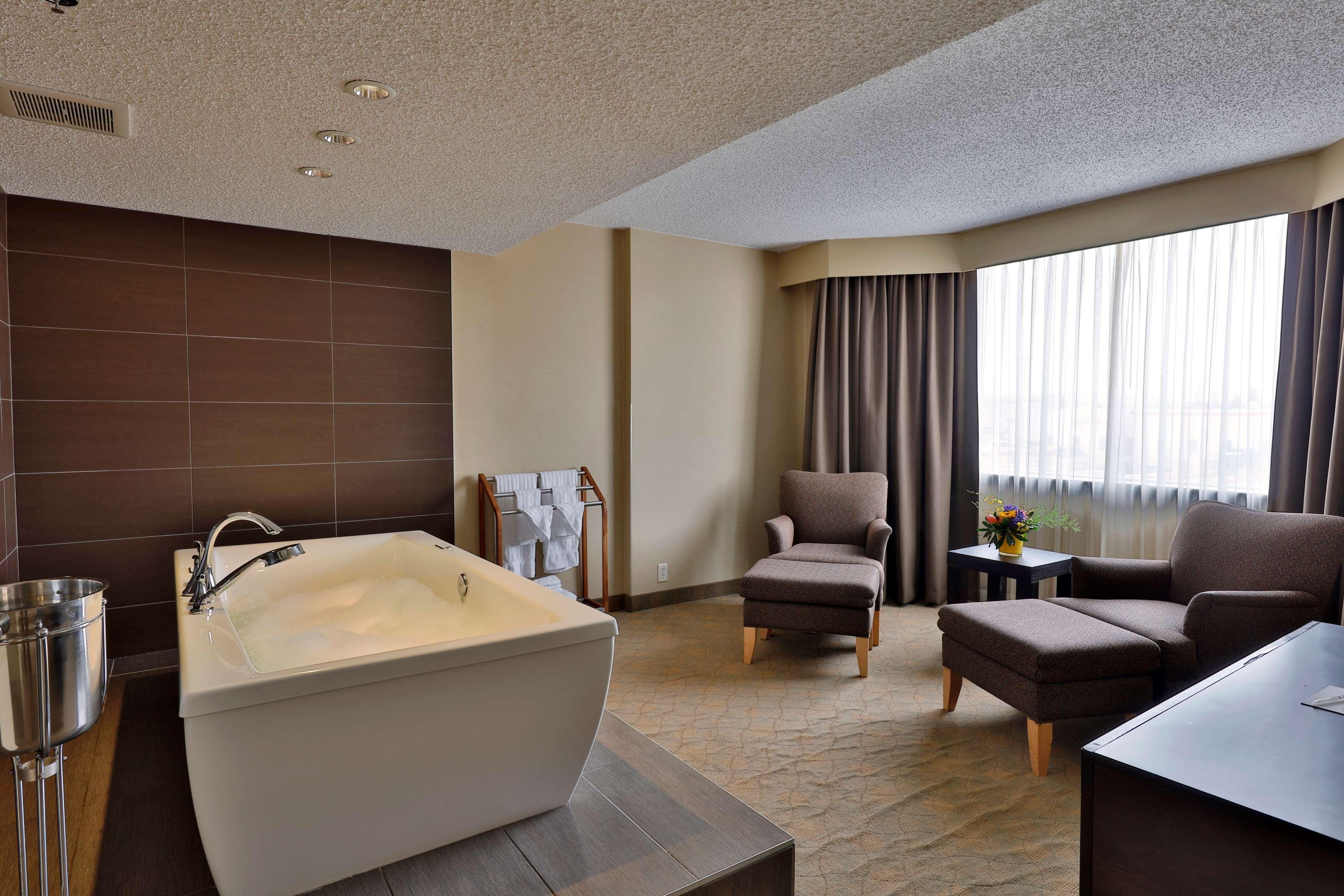 A modern bathroom with a bathtub, a chair, a small table with a vase, and a large window with curtains.