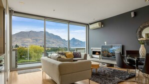 A modern living room with a large window, a fireplace, a flat-screen TV, and a dining area. - Pounamu Terrace | Central · Lakeview · Fireplace (Queenstown)