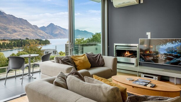 A modern living room with a large TV, a fireplace, and a scenic view of mountains and a lake. - Pounamu Terrace | Central · Lakeview · Fireplace (Queenstown)