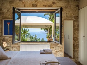 A bedroom with a stone wall, a large window with an open view of the sea, a bed with white linens, and a framed picture on the wall. - Villa Vollard  | 5 Bedrooms with Private Pool Jacuzzi and Panoramic Sea View (Heraklion)