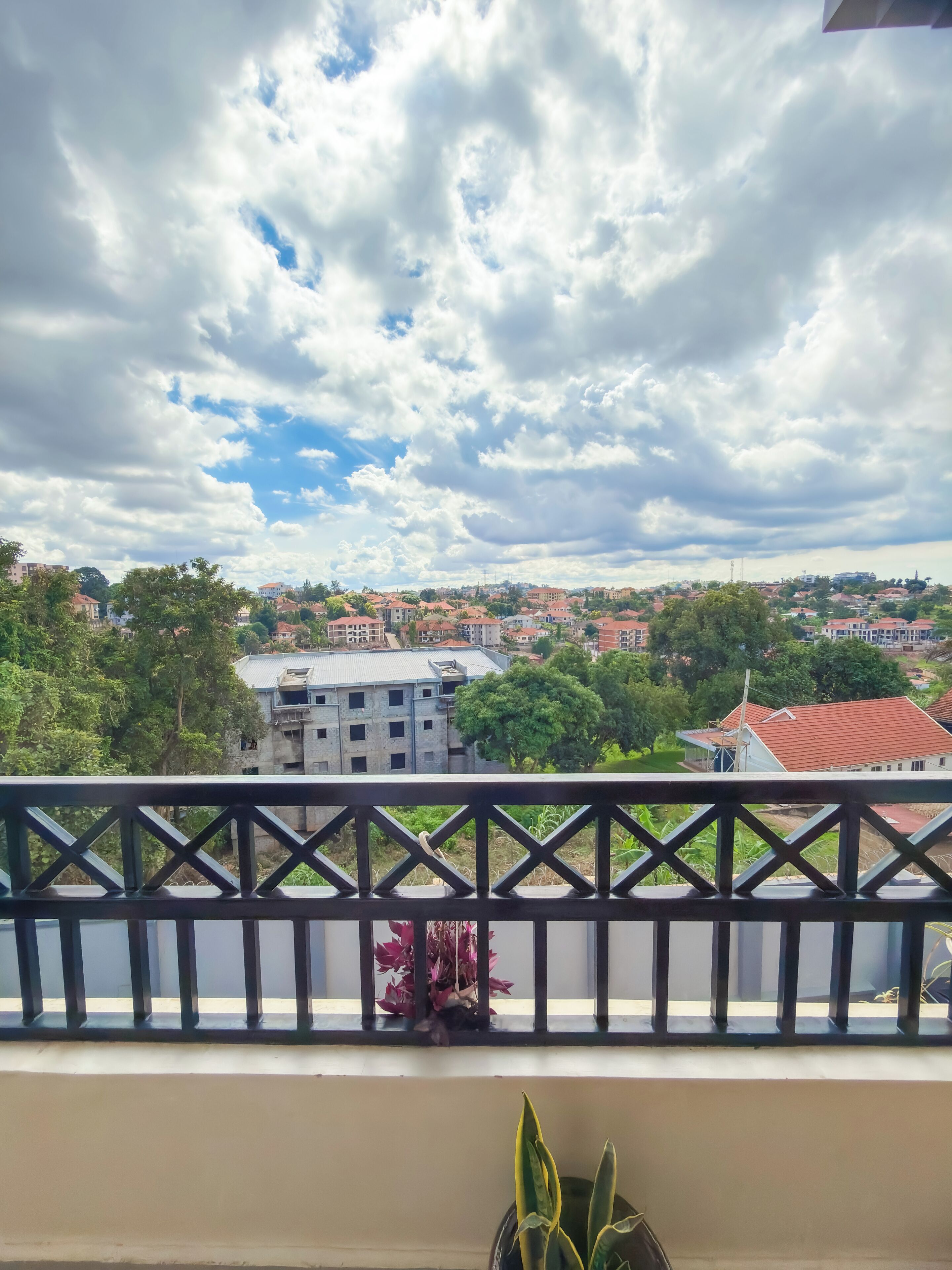 A balcony with a railing, a potted plant, and a view of a town with buildings and trees under a partly cloudy sky.