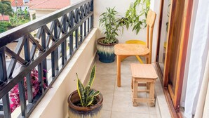 Un balcón con vista a una zona residencial, una silla de madera, una mesita pequeña y dos plantas en macetas.