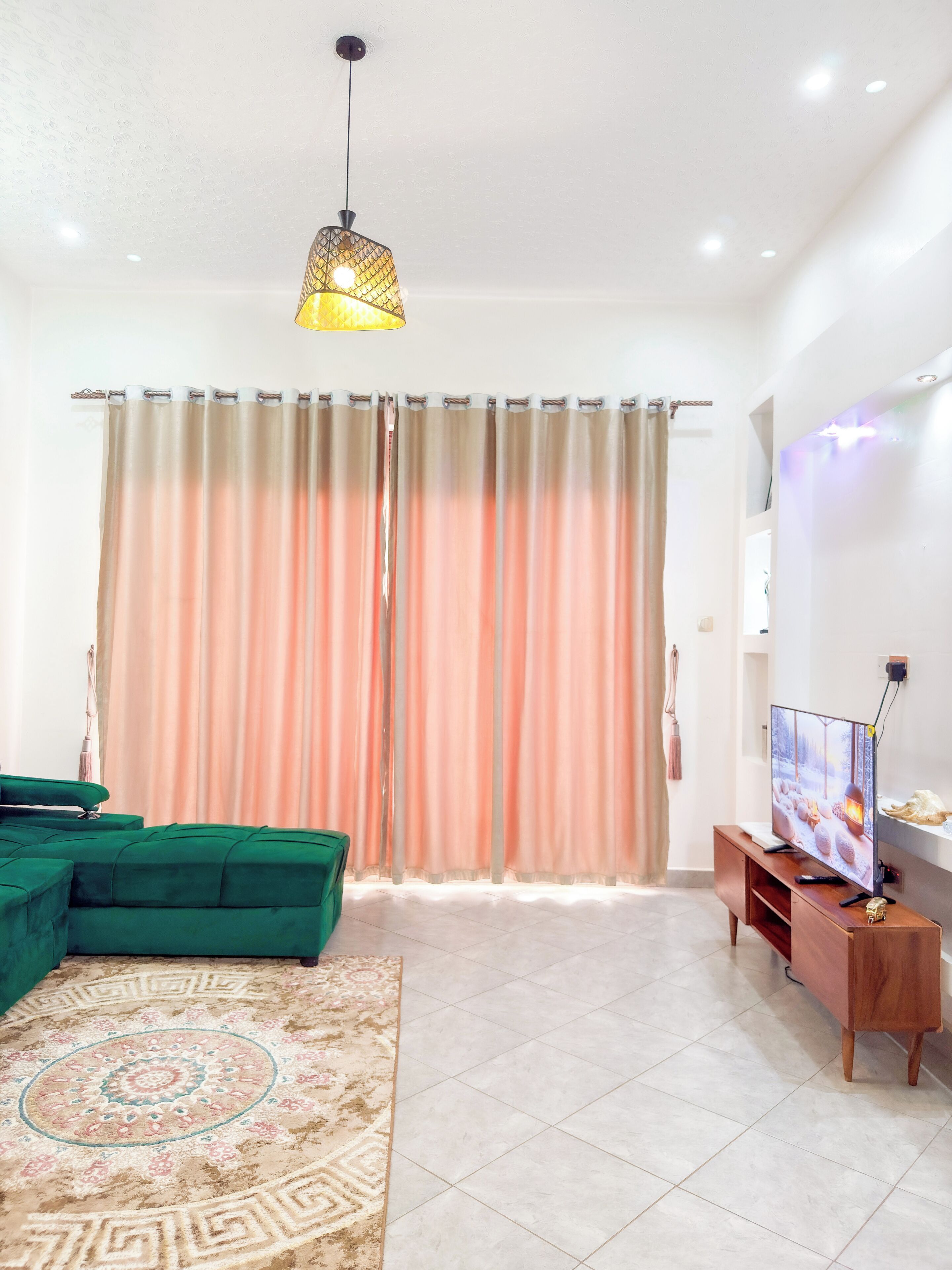 A living room with a green sofa, a patterned rug, a television on a wooden stand, and a ceiling light.