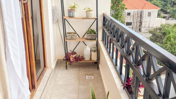Un balcón con barandilla de metal, una planta en maceta, un estante con plantas y vistas a edificios al fondo.