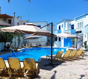 A swimming pool with sun loungers and umbrellas, surrounded by white buildings with blue accents. - Omega Rooms - Studio Room For 2 (Anaxos Skoutarou)