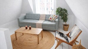 Een loft-achtige kamer met een lichtblauwe bank, een houten bijzettafeltje, een geweven vloerkleed, een plantenpot van riet en een houten stoel met een gestreept kussen.